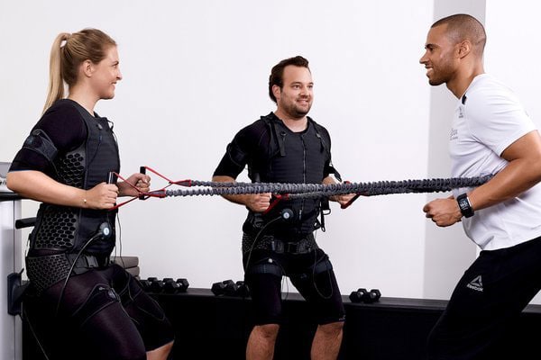Drei Personen trainieren mit Widerstandsbändern, lachen und tragen schwarze Trainingsanzüge.