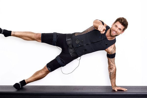 Mann lächelt, balanciert in schwarzem Sportoutfit, sichtbare Tattoos; vor weißem Hintergrund.